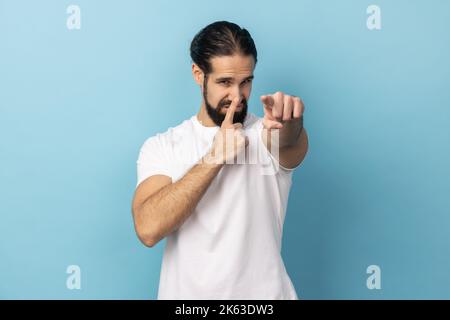 Portrait of man wearing white T-shirt looking with incredulous suspicious gaze and touching nose, gesturing you are liar, suspecting falsehood. Indoor studio shot isolated on blue background. Stock Photo