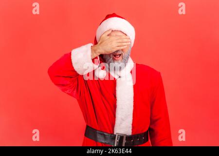 Portrait of elderly man with gray beard wearing santa claus costume covering eyes with palm, does not want to see, avoiding problems. Indoor studio shot isolated on red background. Stock Photo