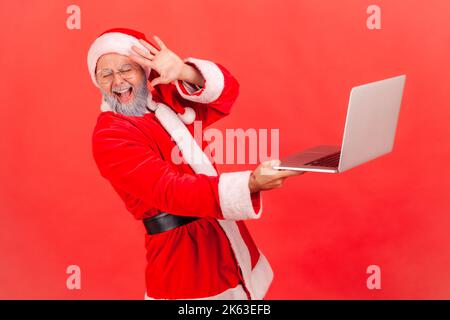 Portrait of elderly man with gray beard wearing santa claus costume standing with computer in hands, sees terrible content on screen, covering face. Indoor studio shot isolated on red background. Stock Photo