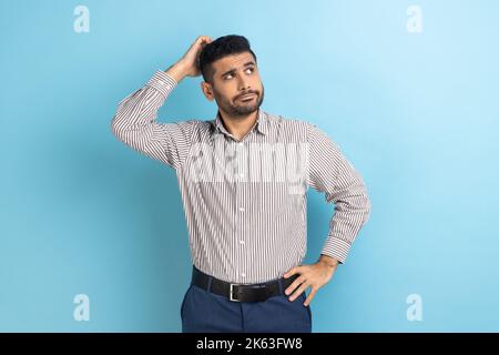 Portrait of young pensive businessman with beard touching head and thinking about important question, making hard decision, wearing striped shirt. Indoor studio shot isolated on blue background. Stock Photo