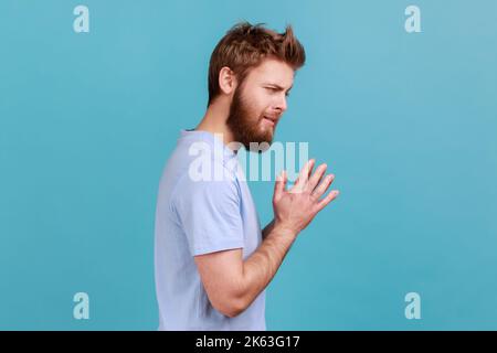 Side view of cunning bearded man schemes something keeps hands together and makes plans, sly expression looking at camera thoughtfully. Indoor studio shot isolated on blue background. Stock Photo