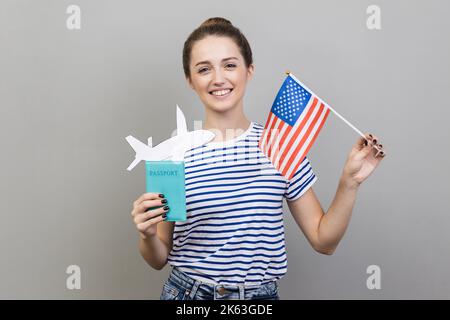 Portrait of smiling woman wearing striped T-shirt standing with American flag and passport with paper plane, travel abroad. Indoor studio shot isolated on gray background. Stock Photo