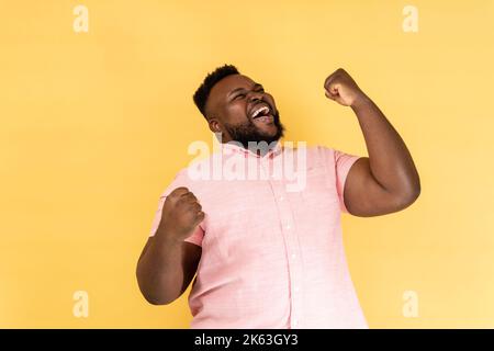 Portrait of extremely happy man wearing pink shirt celebrating his success, standing with clenched fists, screaming happily. Indoor studio shot isolated on yellow background. Stock Photo