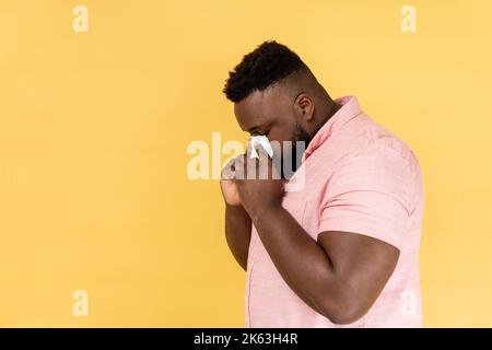 Side view portrait of man wearing pink shirt hiding face in hand with white handkerchief, crying, feeling stressed, worried facial expression. Indoor studio shot isolated on yellow background. Stock Photo