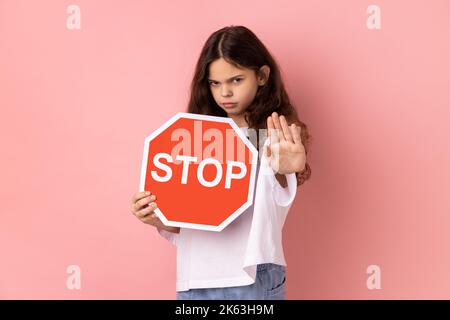 No, prohibited. Strict bossy little girl in T-shirt looking seriously at camera and showing stop gesture, holding road traffic sign as warning of ban. Indoor studio shot isolated on pink background. Stock Photo