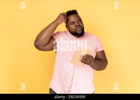 Portrait of thoughtful man wearing pink shirt thinking over smart idea, holding pencil and notebook to write plans, to-do list. Indoor studio shot isolated on yellow background. Stock Photo