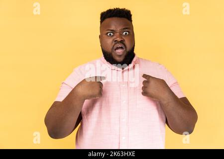 Portrait of bearded astonished man wearing pink shirt pointing at himself, asks who me, has surprised expression, shocked being picked. Indoor studio shot isolated on yellow background. Stock Photo