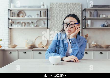 sad and thinking woman in kitchen, depressed asian woman sitting at table at home, young wife in glasses disappointed. Stock Photo