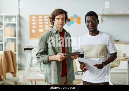 Waist up portrait of two young IT professionals looking at camera while standing in office setting and holding documents Stock Photo