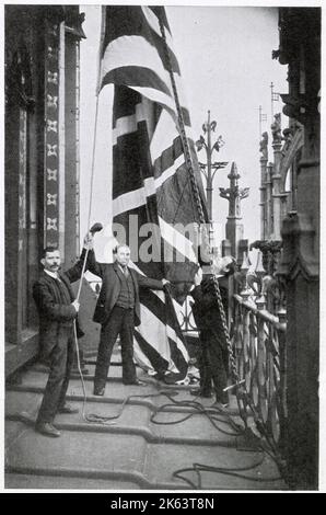 When the Sovereign is in Houses of Parliament building the Royal Standard is flown, on Victoria Tower. Photograph showing two men hoisting the Union Jack flag. Stock Photo
