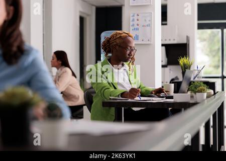 Employee working on laptop with financial report in business office open space, woman sitting at workplace desk. Young woman thinking, looking at documents side view medium shot Stock Photo