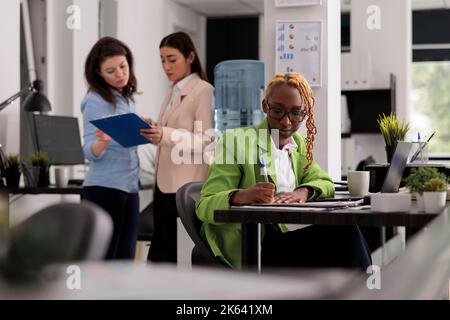 Diverse employees in coworking open space, worker writing on clipboard, sitting at workplace desk. Young african american woman working with company documents, taking notes Stock Photo