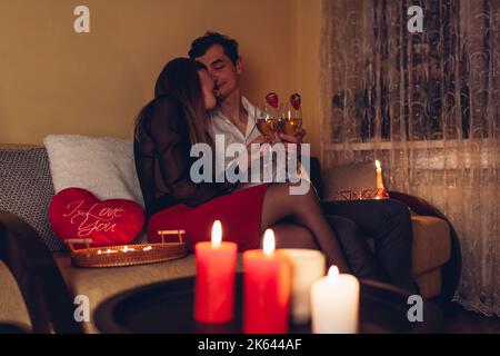 Young Valentines Day couple in love clinking glasses drinking wine with strawberries having romantic dinner with candles celebrating at home at night Stock Photo