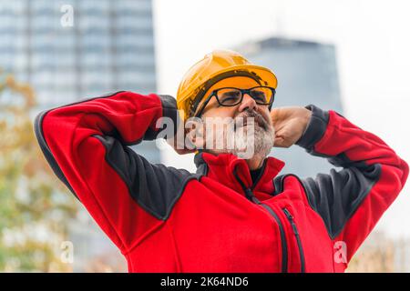 Male caucasian elderly construction worker with gray facial hair wearing bright red fleece and yellow helmet, looking up in the sky and holding the back of his helmet with two hands. Outdoor medium closeup shot. High quality photo Stock Photo
