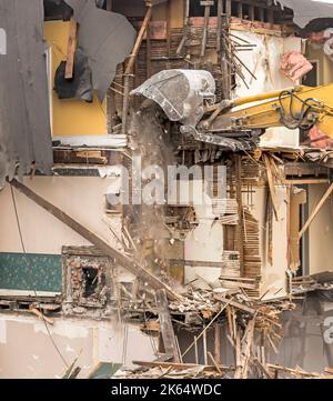 Demolishing a building with a large backhoe. Air is dusty. Bricks are falling from the bucket. Wall slats, wallpaper, insulation, and more is visible. Stock Photo