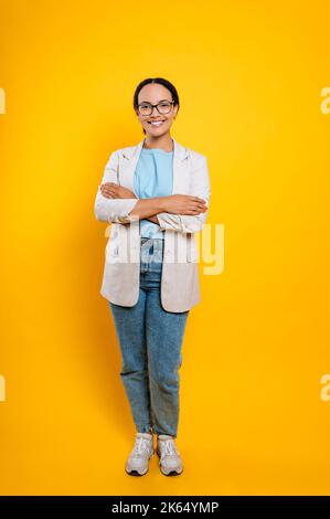Full length photo of satisfied successful pretty, hispanic or brazilian business woman, company employee, stylishly dressed, with glasses, stand on isolated orange background with arms crossed, smiles Stock Photo