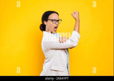 Amazed excited confident strong young brunette hispanic or brazilian woman, business lady, showing biceps muscles, demonstrates her own strength, looks at camera, stand on isolated orange background Stock Photo