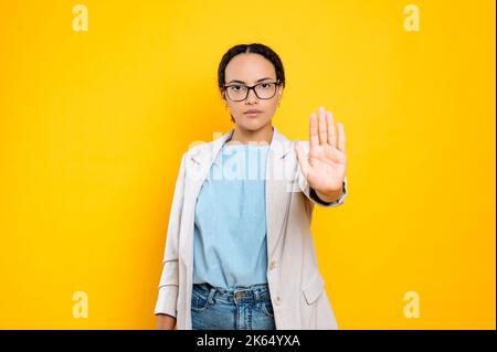 Serious concerned resolute young brunette hispanic or brazilian woman, showing stop gesture with palm, looks at camera, stand on isolated orange background. Against discrimination on women Stock Photo