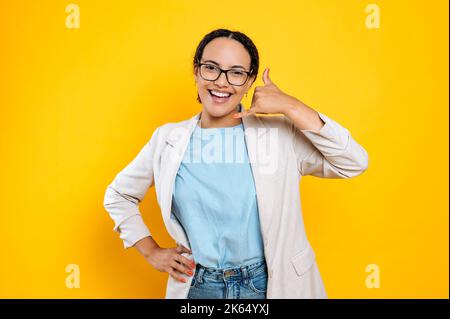 Cheerful friendly pretty hispanic or brazilian young woman, stylishly dressed, with glasses, doing phone gesture with hand, like says call me back, smiles at camera,stand on isolated orange background Stock Photo