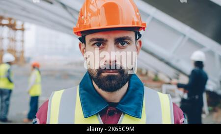 Close-upportrait of serious guy wearing safety helmet and west standing in construction site with group of people working in background Stock Photo