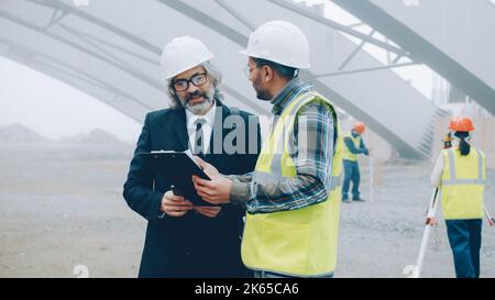 Project manager is talking to builder discussing construction work in buiding area and using tablet. People and business communication concept. Stock Photo