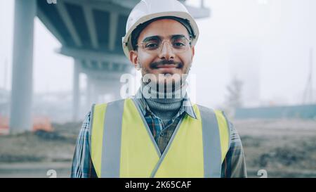 portrait of cheerful Arab man wearing safety helmet and vest smiling standing against urban construction background alone and looking at camera Stock Photo