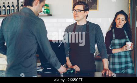 Handsome male cashier is accepting contactless smartphone payments while attractive young waitress is bringing takeaway coffee to customers in modern coffee-shop. Stock Photo