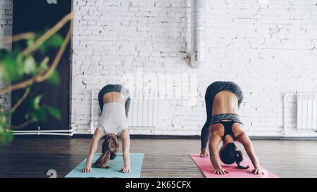 Young blond girl is enjoying individual yoga practice with friendly female instructor in light studio. Women are doing sequence of asanas on bright mats. Friendly relaxing atmosphere. Stock Photo