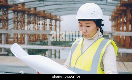 portrait of young Asian forewoman wearing helmet studying blueprint in construction site. People and occupation concept. Stock Photo