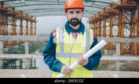 Portrait of male builder wearing helmet and vest standing outside construction site holding blueprint and looking at camera. People and profession concept. Stock Photo