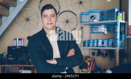 Portrait of confident man bike repair workshop owner standing in his workplace and looking at camera. Bicycle spare parts, reparing equipment and tools are in background. Stock Photo