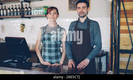 Portrait of two confident waiters in aprons standing at cashier's desk in new coffee-house and smiling. Successful business, happy people and food service concept. Stock Photo