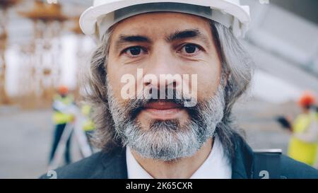 Close-upportrait of mature businessman standing at industrial site wearing helmet and looking at camera. Investors and building concept. Stock Photo