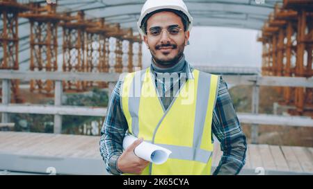 Portrait of good-looking Middle Eastern guy wearing safety helmet and holding construction plan outdoors. Employees and business concept. Stock Photo