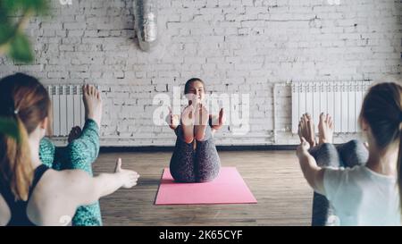 Pretty female yoga instructor is demonstrating different variations of boat pose, talking to students and laughing. Friendly relaxing atmosphere in class. Fun in yoga studio concept. Stock Photo