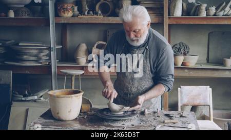 Creative male sculptor is making unusual broad bowl while standing at working table in his workshop. Bearded senior man is liiking at his clayware and appraising design and quality. Stock Photo