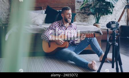 Young handsome musician popular blogger is recording video with camera playing the guitar and singing song sitting on floor in bedroom. Blogging and music concept. Stock Photo