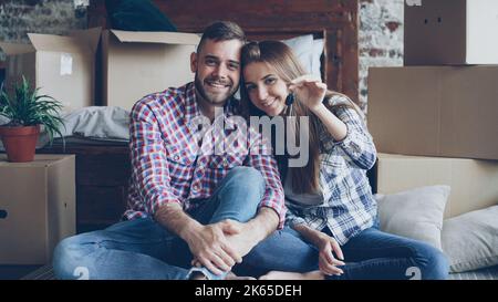 Portrait of happy young couple sitting together in new flat, holding keys and looking at camera. Nice loft style interior, carton boxes and double bed are visible. Stock Photo