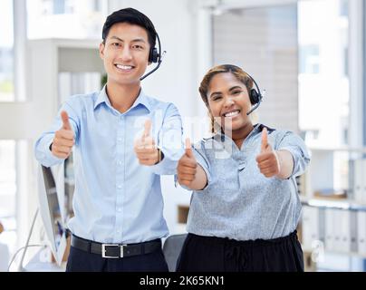 Two young happy male and female call center agents showing thumbs up while answering calls working in an office at work. Pleased asian man and mixed Stock Photo
