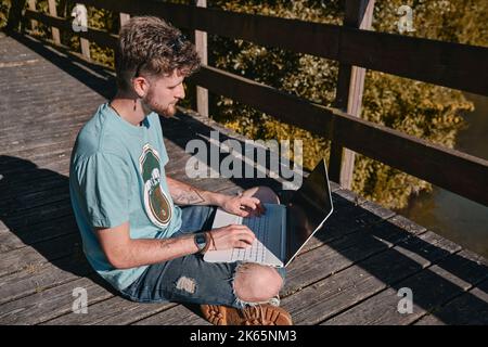 A young tattooed entrepreneur working outdoors on his laptop with a serious face sitting on a bridge Stock Photo