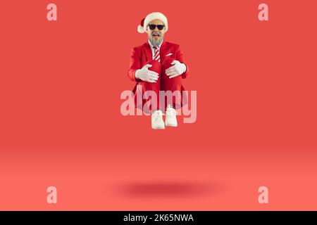 Old cheerful grey-haired man wearing Santa style is sitting levitating in air on red background. Stock Photo
