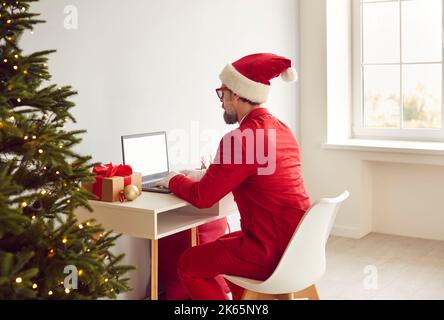 Man in Christmas Santa hat working on laptop while sitting at desk in his office Stock Photo