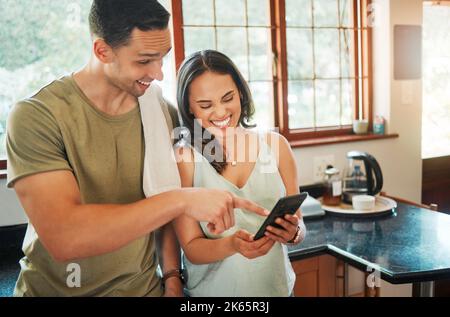 Young cheerful mixed race couple using social media on a phone together in kitchen at home. Hispanic girlfriend showing her boyfriend a funny video Stock Photo