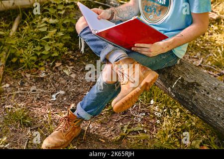 A male student studying with a notebook sitting cross-legged on a log of wood wearing boots Stock Photo
