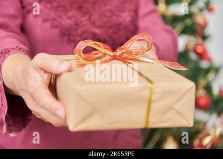 Female hands holding a Christmas gift box wrapped with a decorative bow, handing it towards the camera. Close up composition. Stock Photo