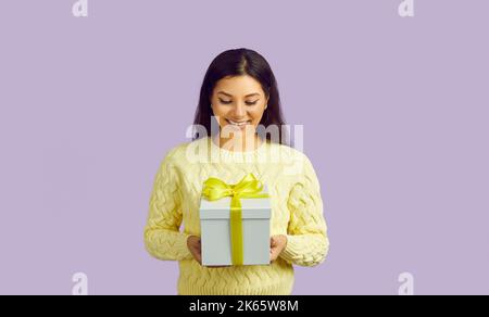 Joyful smiling young woman on pastel purple background holding festive gift box. Stock Photo