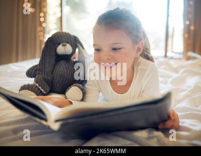 Happy smiling little girl lying on a bed at home and reading a childrens book to her toy teddy. Cute child relaxing and playfully reading a story to Stock Photo