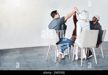 Diverse group of businesspeople giving each other a high five sitting on chairs in a meeting in an office at work. Happy women and men joining their Stock Photo