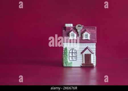 A small ceramic house with a key hanging from the roof on a background of red painted wood texture and copy space. Real state concept. Stock Photo