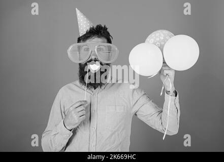 Hipster in giant sunglasses celebrating. Guy in party hat with air balloons celebrates. Man with beard on cheerful face holds smiling mouth on stick Stock Photo